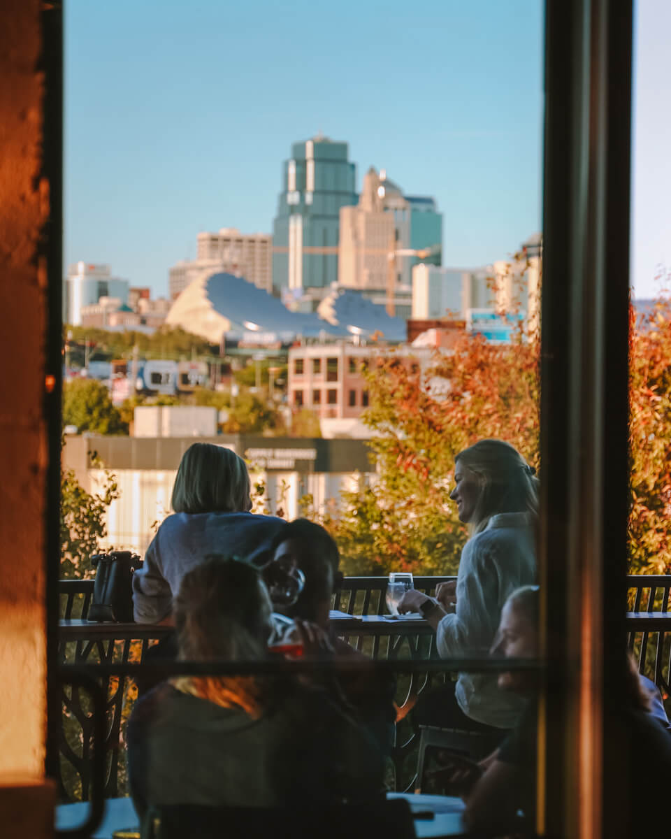 Several people are seated at an outdoor table on a sunny day, engaging in conversation and enjoying drinks. In the background, a city skyline is visible, with modern and tall buildings under a clear blue sky. Trees with autumn foliage surround the area.