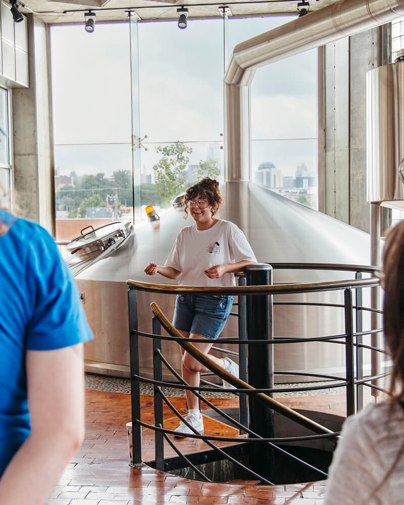 A person stands on an indoor spiral staircase with metal railings, smiling outdoors into a large window where distant city buildings are visible. The person is wearing a white T-shirt, blue shorts, and sneakers. Brewing equipment is seen behind them.