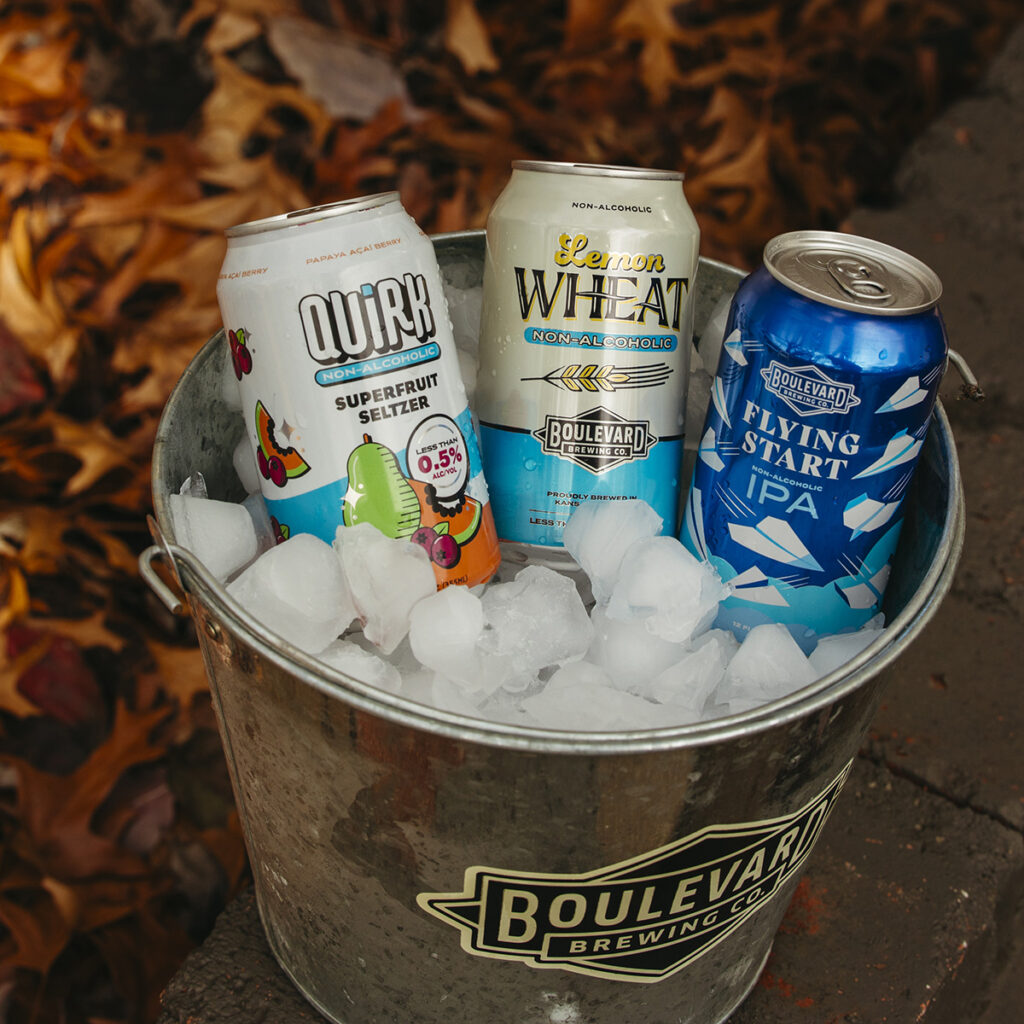 A metal bucket filled with ice holds three beverage cans: Quirk Superfruit Seltzer, Boulevard Lemon Ginger Radler, and Boulevard Flying Start IPA. The bucket with a Boulevard Brewing Co. logo is placed outdoors amid autumn leaves.