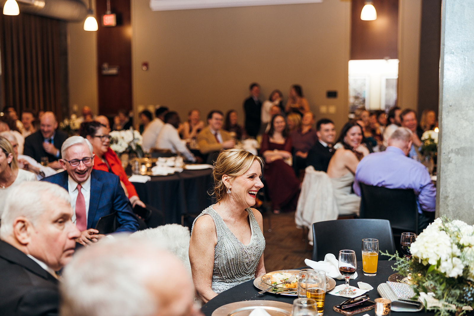 A woman in a silver dress laughs while sitting at a table during a formal event. Around her, guests are seated at tables decorated with white flowers. The room is filled with people in formal attire, and there are drinks and food on the tables.