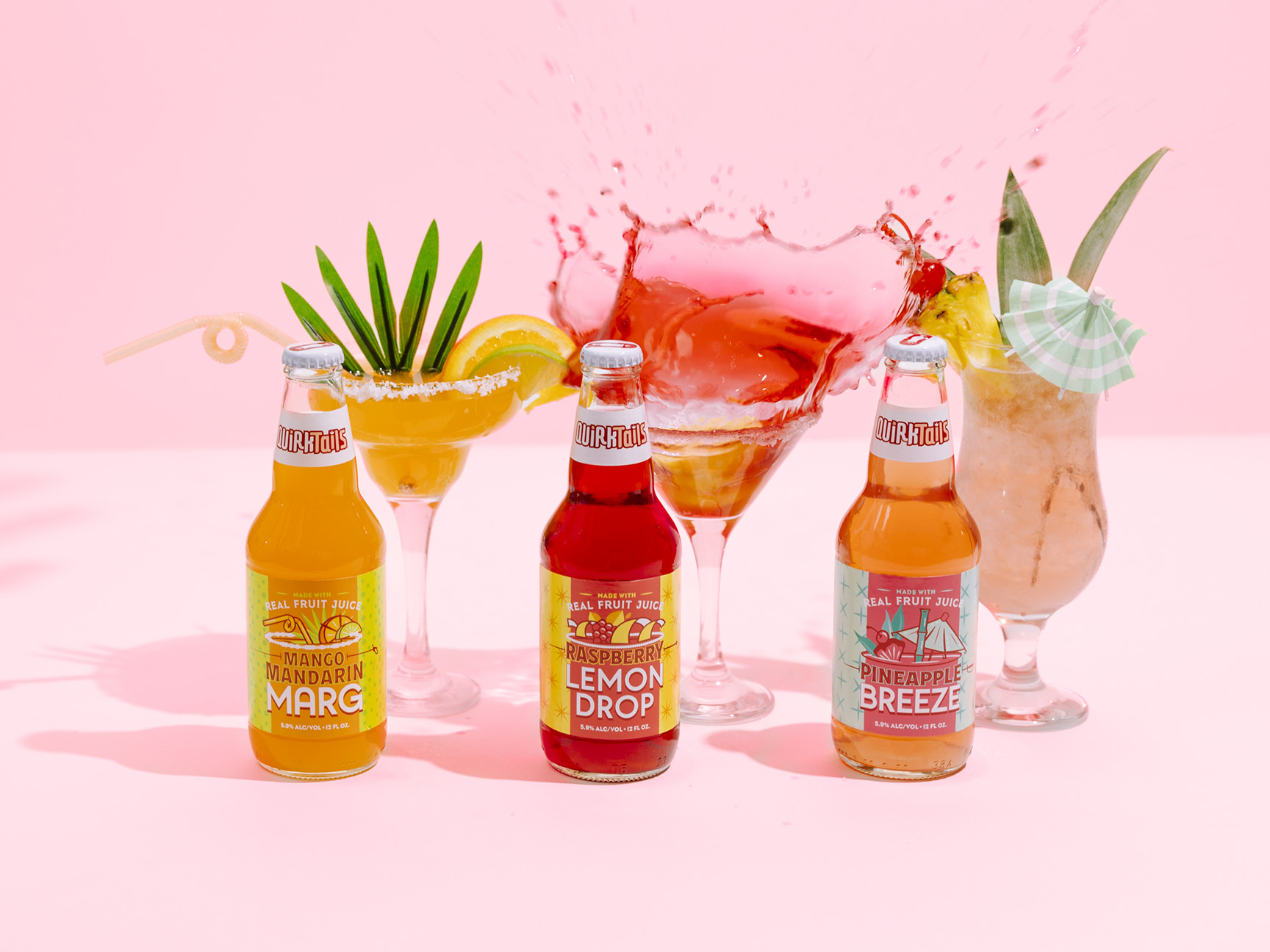 Four bottled beverages are displayed in front of colorful cocktails, set against a pink background. From left to right, the bottles are labeled Mango Mandarin Marg, Raspberry Lemon Drop, and Pineapple Breeze. One cocktail splashes dramatically in the background.