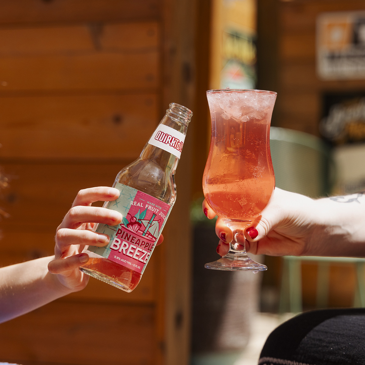 Two hands holding drinks in a toast. The left hand holds a bottle labeled "DURANGO Pineapple Breeze," and the right hand holds a tall glass with a pink beverage. The background shows a wooden structure and a partial view of a chair. Both hands have red nail polish.