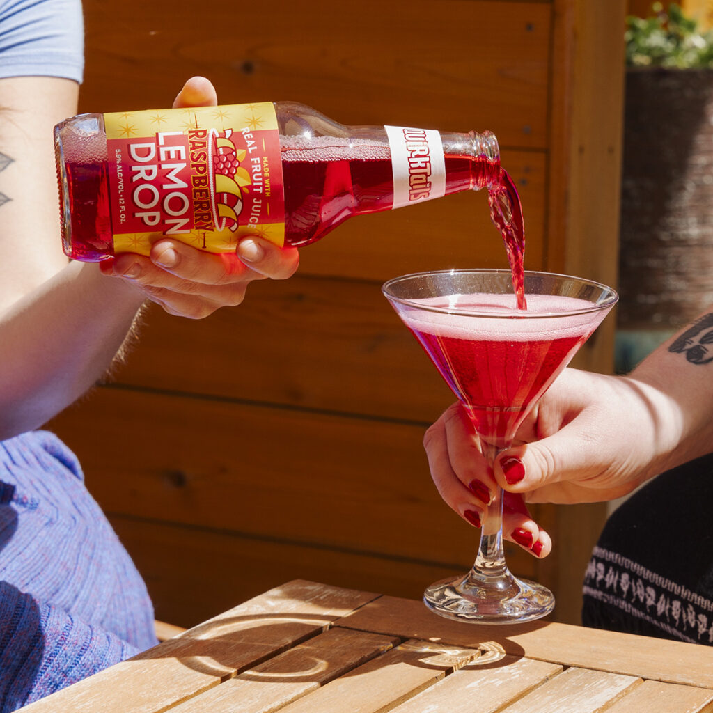 A person pours a bright red drink from a bottle labeled "Raspberry Lemon Drop" into a martini glass held by another person. The scene is set outdoors on a wooden table, with partial views of the people. A corkscrew lies on the table.