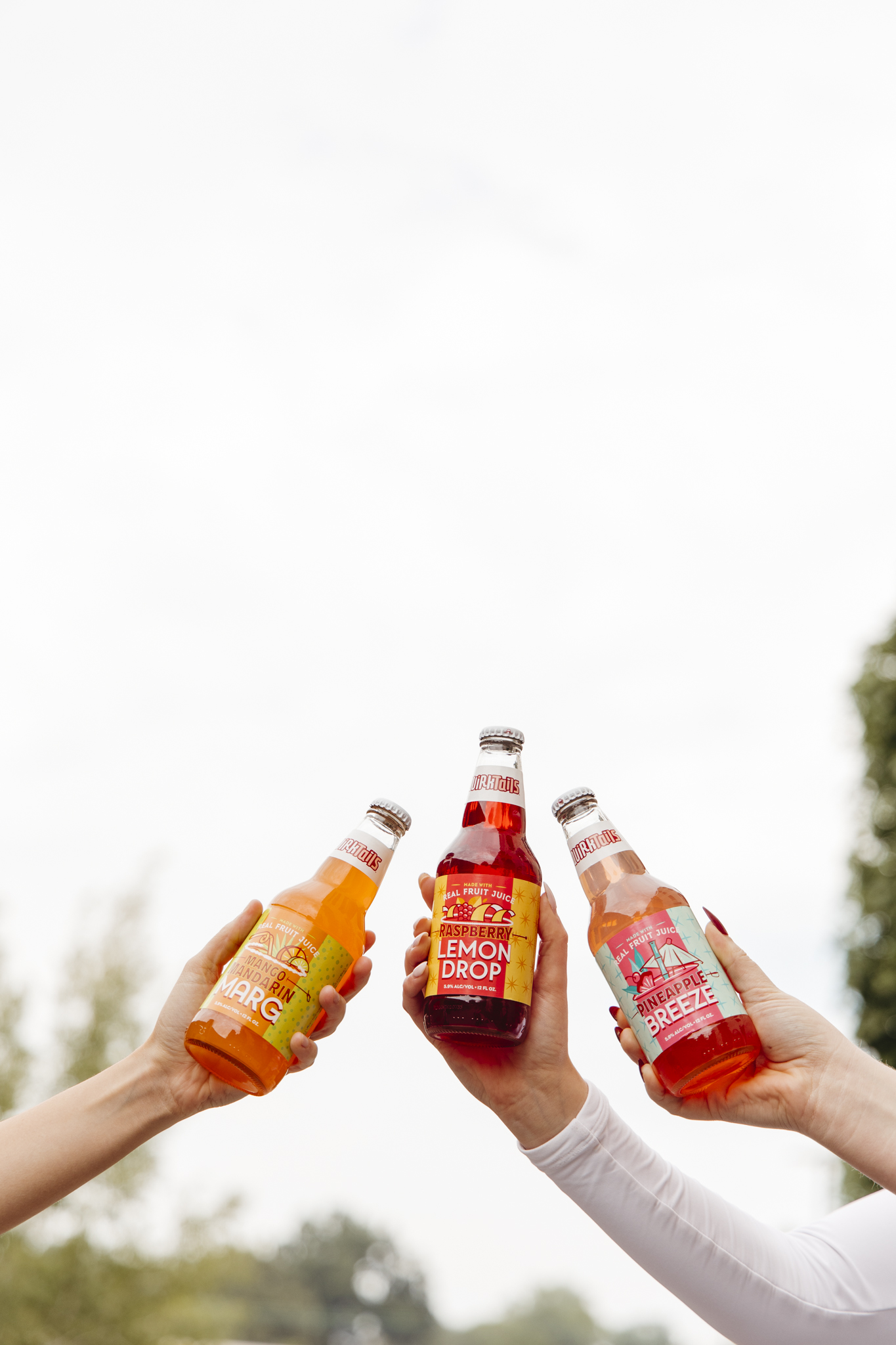 Three hands each holding a different colorful bottled beverage, raised together as if cheering. The labels on the bottles read "Marg", "Lemon Drop", and "Breezer". The background is a bright, outdoor setting with trees visible in the distance.