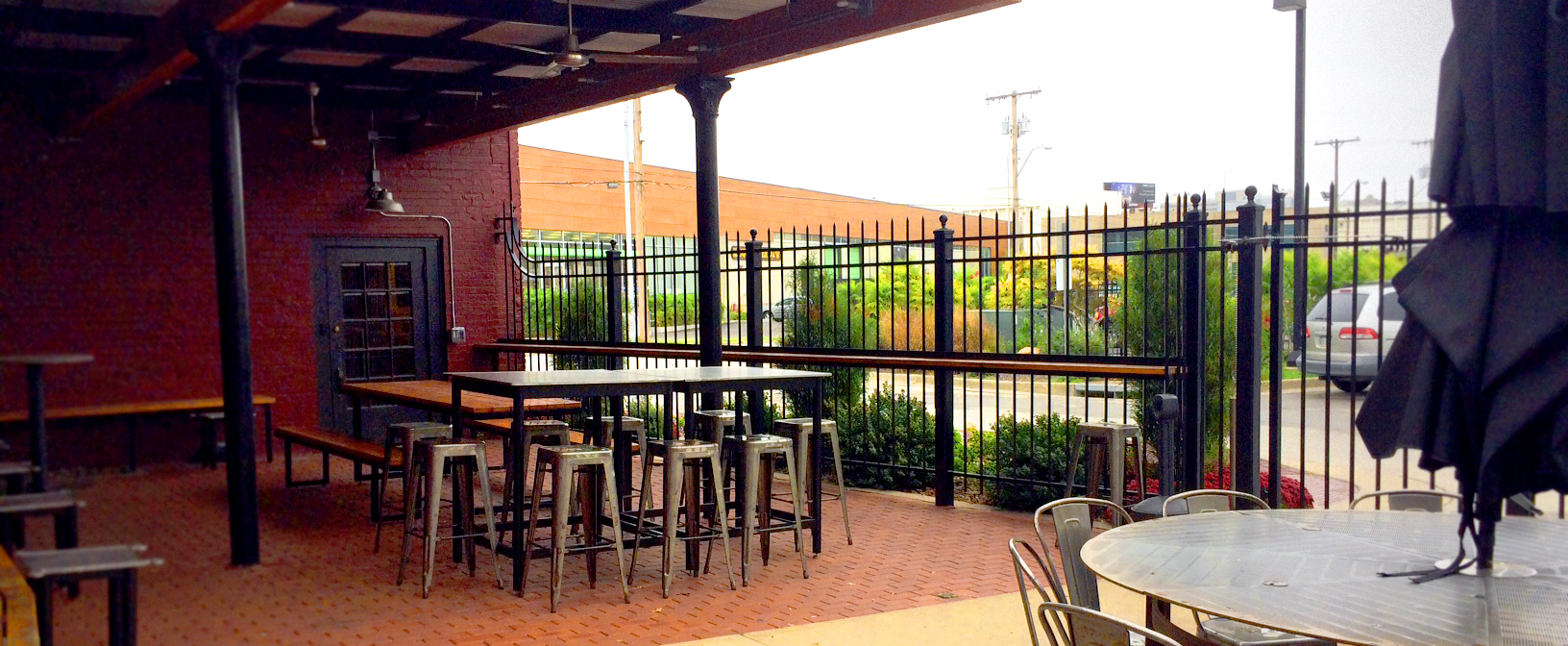 A covered outdoor patio with several high metal tables and stools on a brick-paved floor. The area is fenced with a black metal fence and overlooks a street with parked cars and industrial buildings. There is also an umbrella partially visible on the right side.