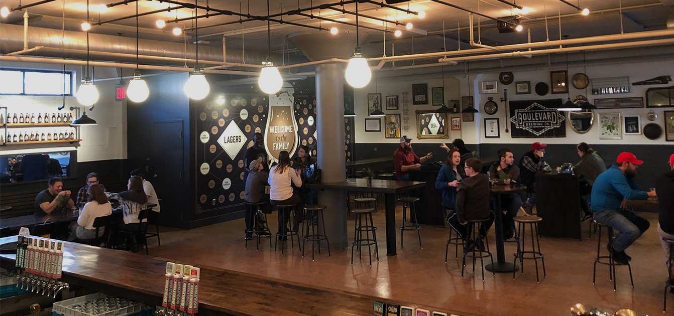 A spacious brewery taproom with patrons seated at high tables enjoying drinks. Various decorations adorn the walls, including signs for lagers, ales, and a logo. Warm lighting hangs from the ceiling, creating a cozy atmosphere. A bar counter is visible in the foreground.