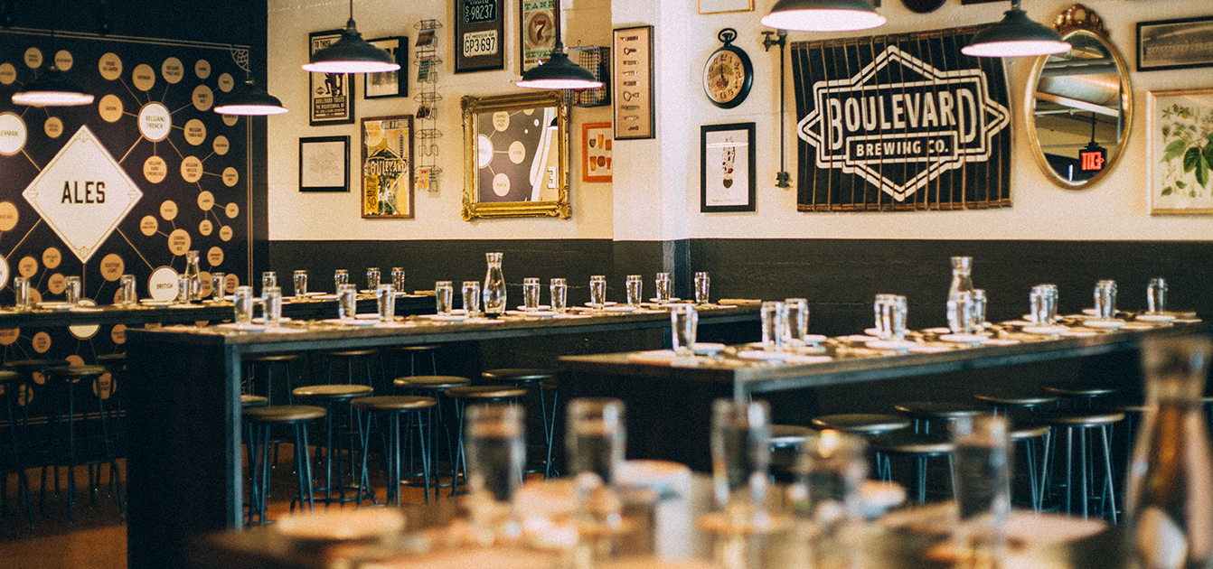 A cozy restaurant with multiple wooden tables and stools, each table set with glass cups and cutlery. The walls are adorned with various signs, including one for "Boulevard Brewing Co," and other framed pictures and artworks under hanging industrial lights.