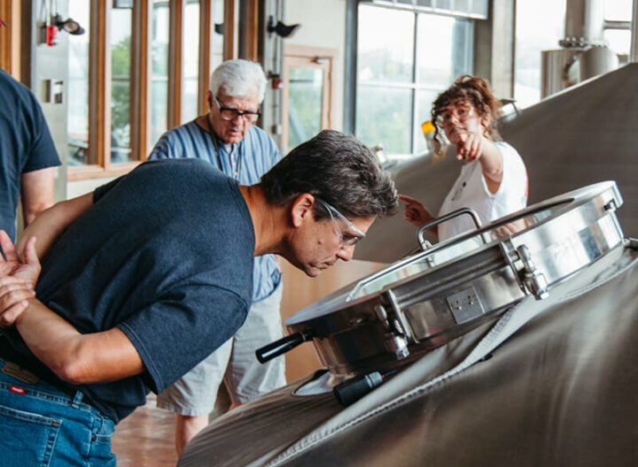 Several people are observing a large industrial machine in a spacious workshop with large windows. One person closely examines the machine while others stand nearby, one of whom is pointing towards something. The floor is tiled, and there are wooden and metal elements in the room.