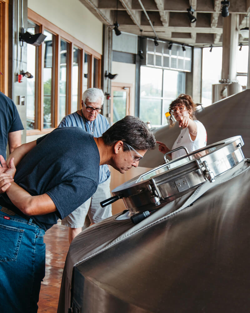 Several people are observing a large industrial machine in a spacious workshop with large windows. One person closely examines the machine while others stand nearby, one of whom is pointing towards something. The floor is tiled, and there are wooden and metal elements in the room.