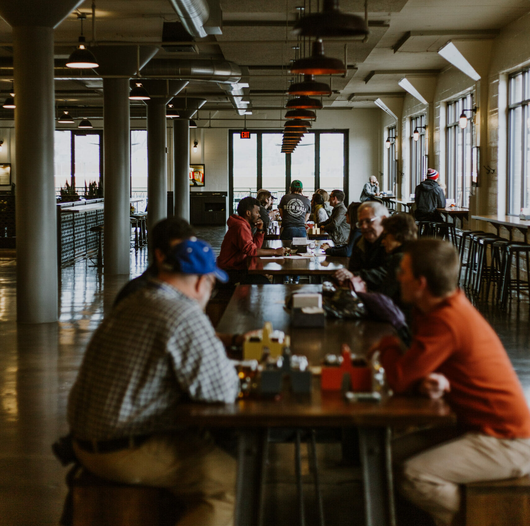 Inside the Boulevard Brewing Co. Beer Hall in Kansas City, people are seated at long wooden tables beneath hanging lights. Some enjoy their meals while others chat, fostering a lively atmosphere. Large windows line the right side, flooding the Tours and Rec Center with natural light.