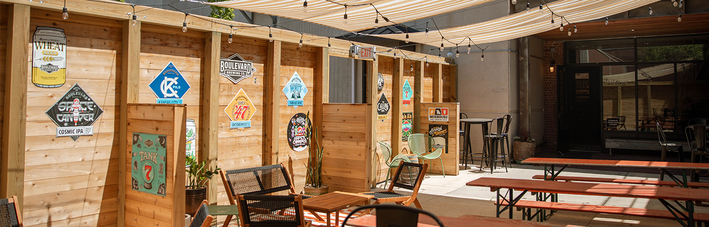 An outdoor patio with wooden walls adorned with colorful vintage signs. String lights hang overhead under striped canopy shades. The seating area includes several tables and benches along with wicker chairs. The background features a door leading inside.