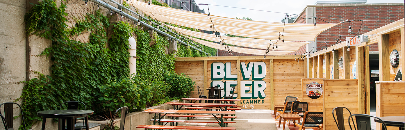 An outdoor seating area with wooden tables and benches, surrounded by green ivy-covered walls. A sign on the wooden wall reads "BLVD BEER." String lights are hung above, and a shade canopy provides cover. A tall building and a smaller brick structure are in the background.