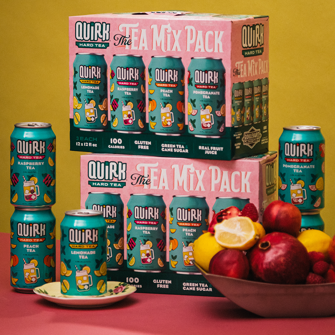 A display of Quirk Hard Seltzer and Hard Tea cans and boxes, labeled "The Tea Mix Pack." Several cans are stacked with visible flavors like peach and raspberry. In the foreground, a bowl of fruits including lemons and pomegranates sits against a yellow background.