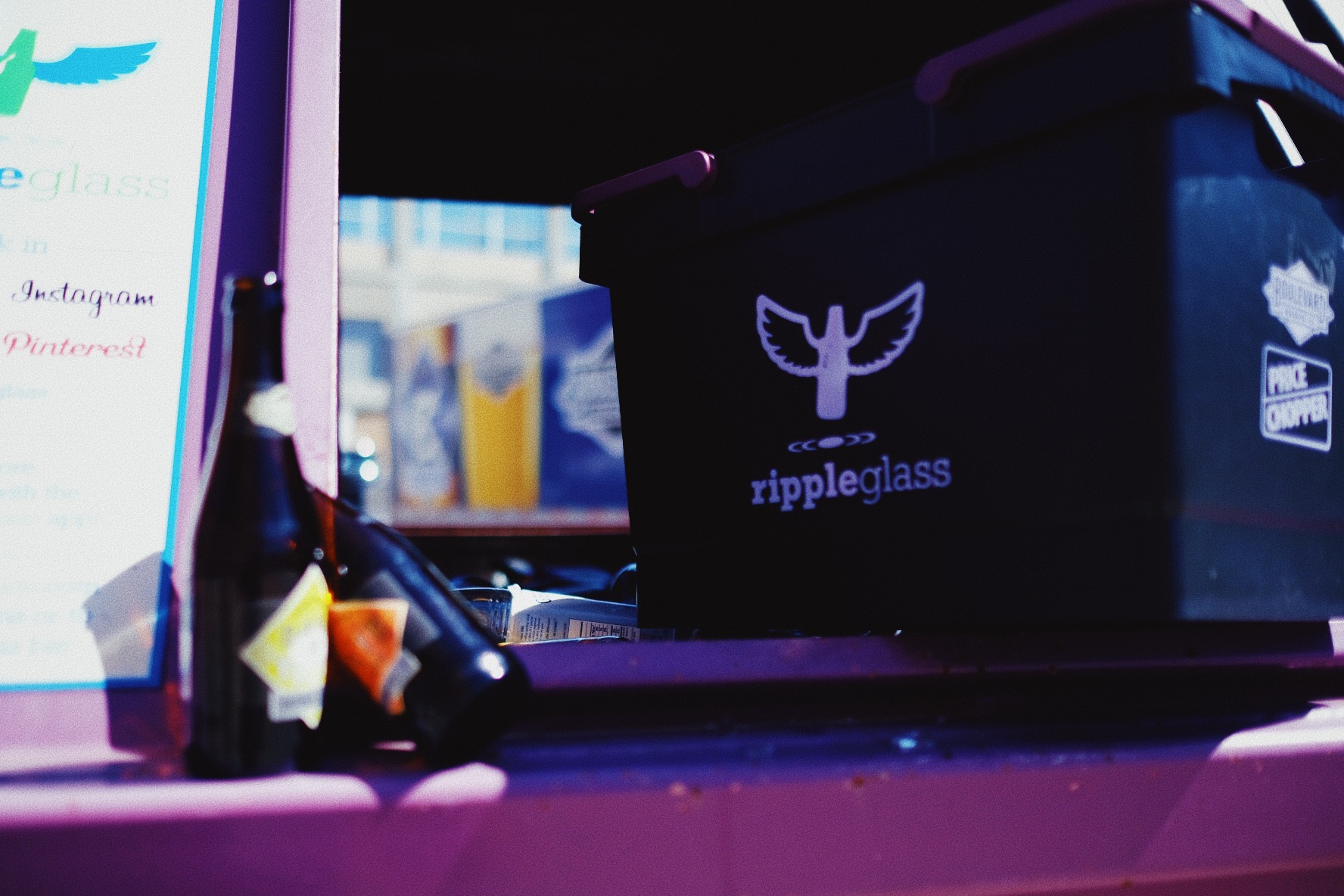 A close-up of a purple recycling bin labeled "rippleglass" with a winged logo, next to a window. A dark glass bottle is placed on the windowsill. The background is blurred, with some text partially visible on the left.