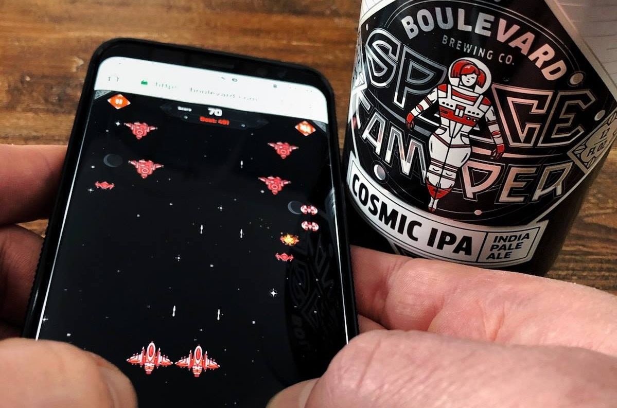 A person plays a retro space shooter game on a smartphone, with red spaceships and a score display. Next to the phone is a can of Boulevard Brewing Co.'s Space Camper Cosmic IPA. The can features a futuristic astronaut design.