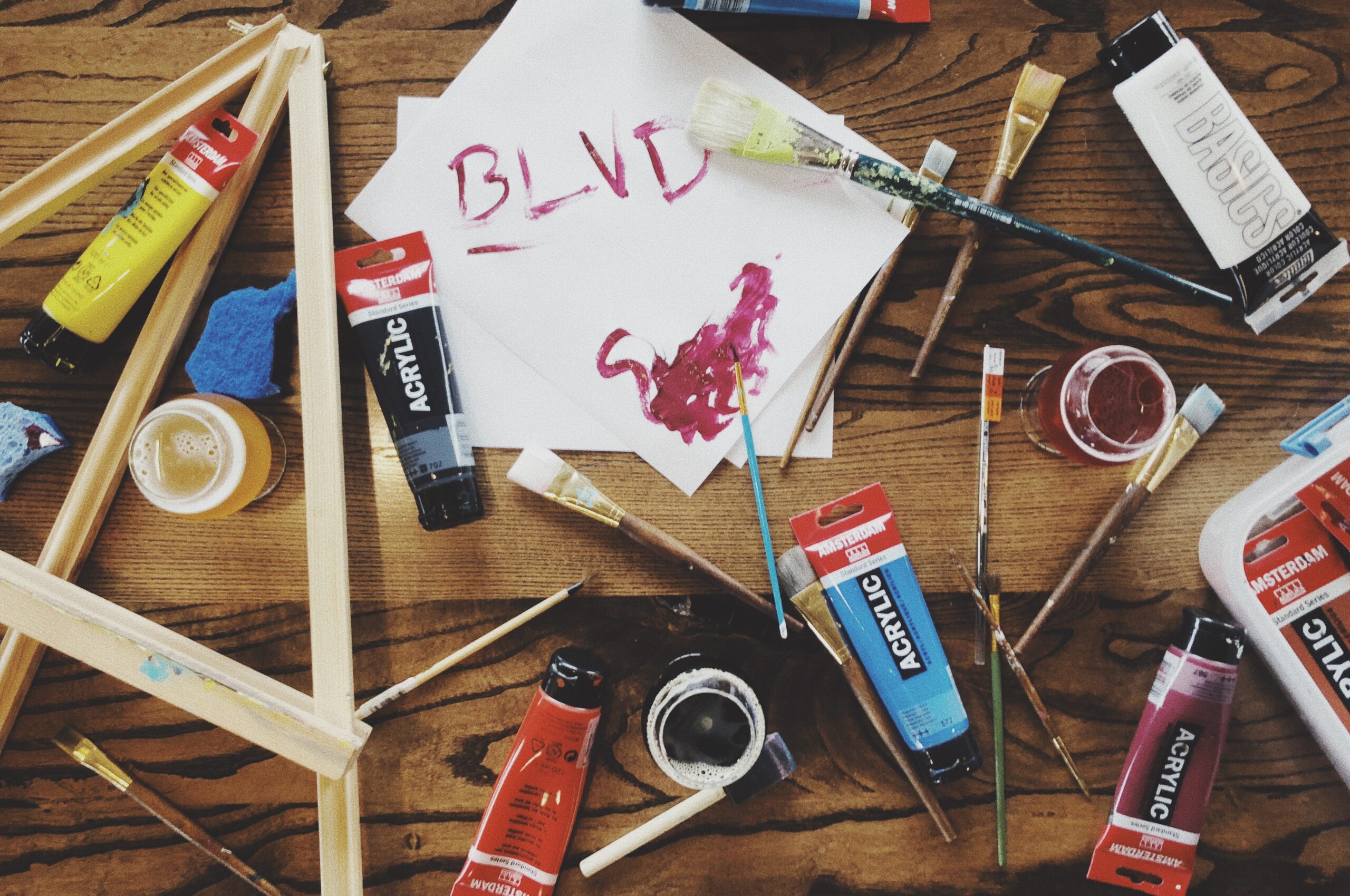 A wooden table scattered with various acrylic paint tubes, brushes, and painting supplies. A sheet of paper with "BLVD" written in red paint sits in the center, alongside a few paint splatters. A wooden frame and two cups are also visible.