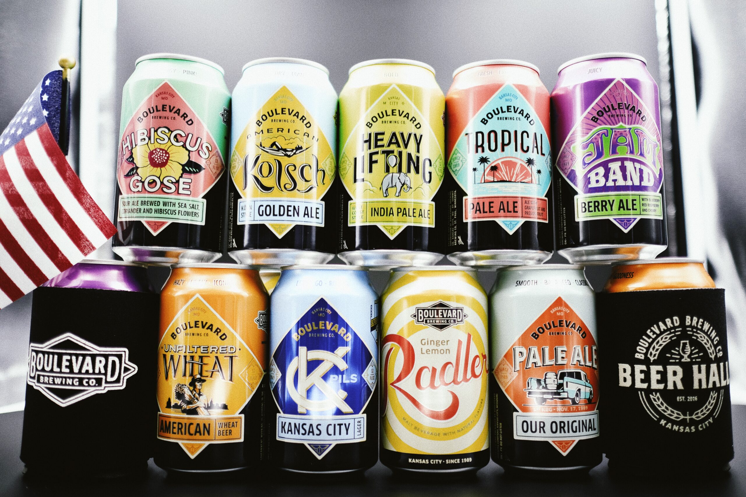 A display of assorted Boulevard Brewing Co. beer cans in various colors and designs. They are arranged in two rows with an American flag to the left and a black can holder to the right.