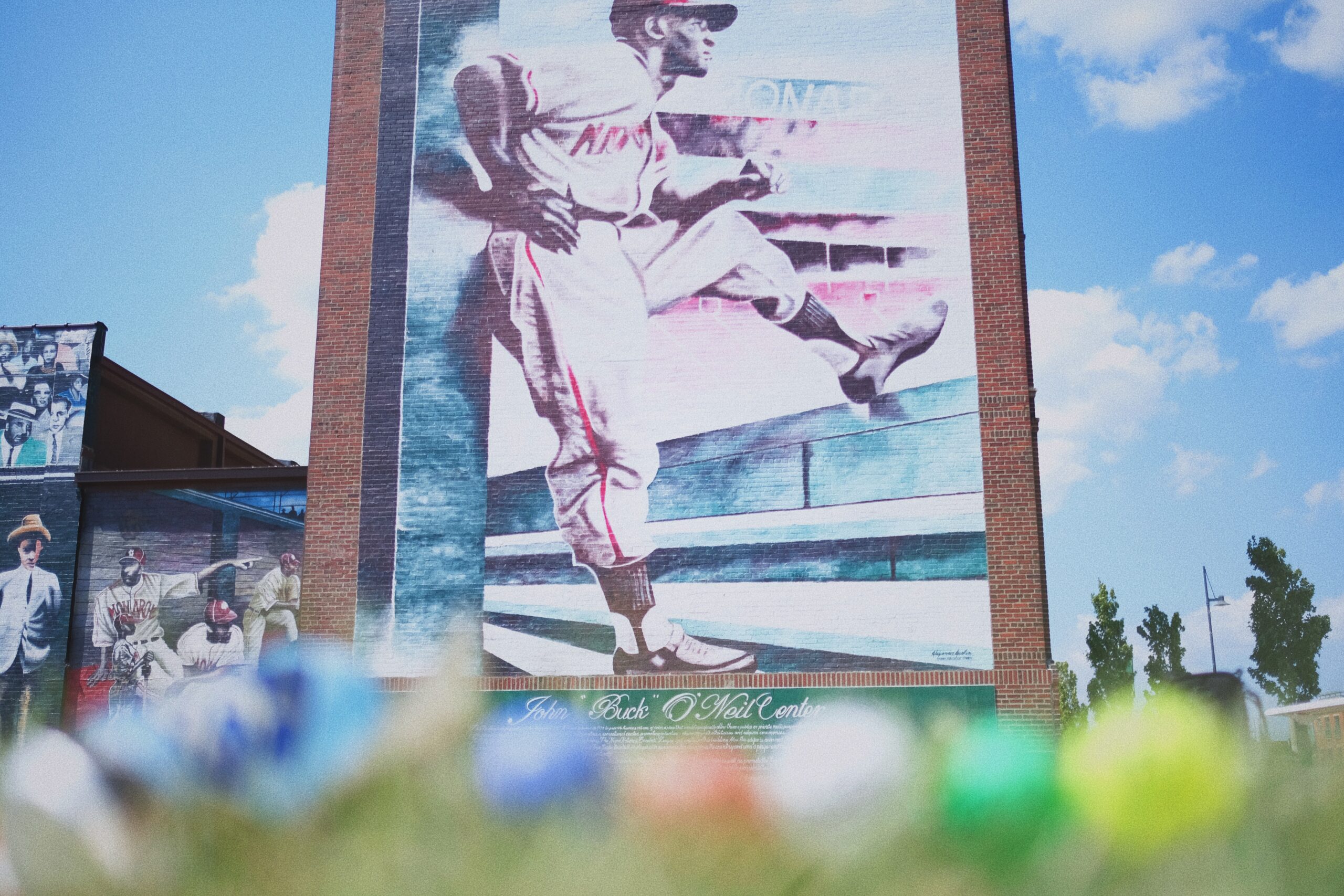 A vibrant mural on a brick building depicts a baseball player in mid-pitch. The artwork is surrounded by a clear blue sky, with blurred colorful objects in the foreground.