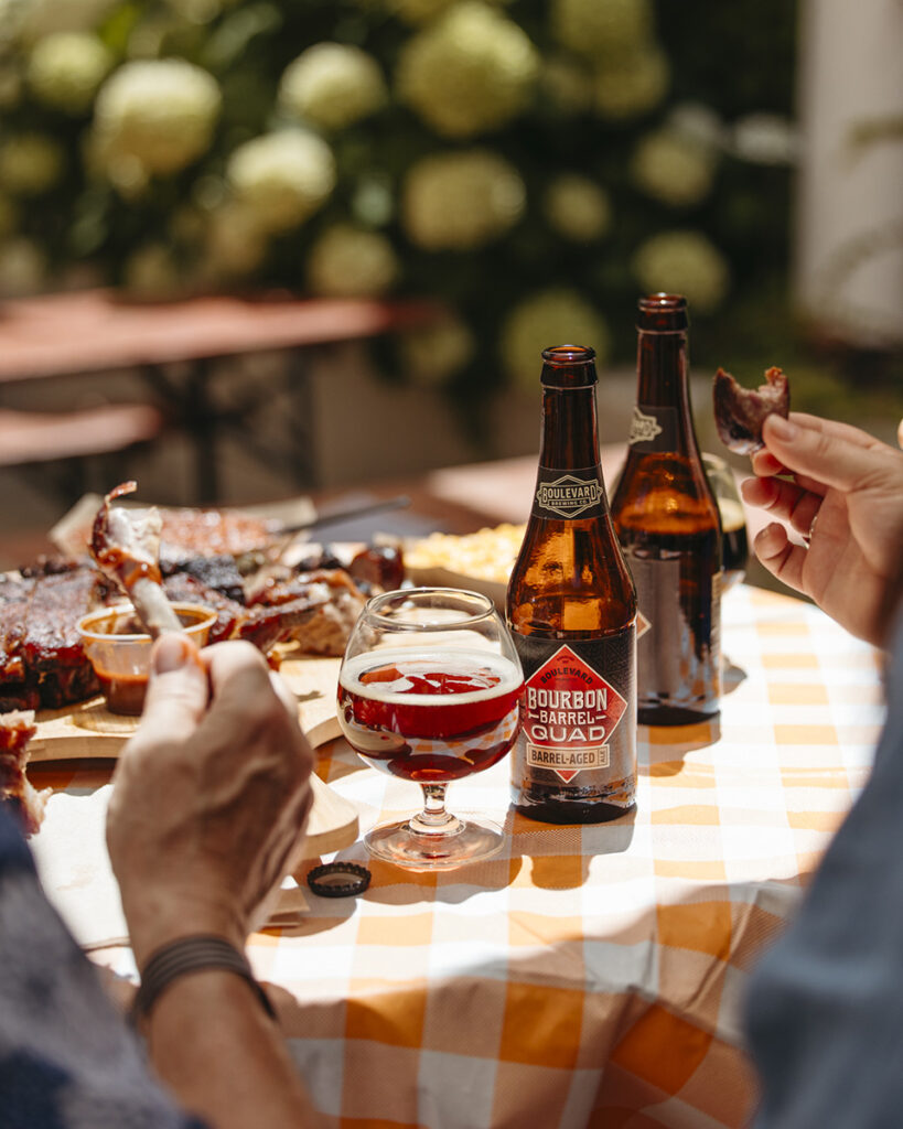 Outdoor table set with two Bourbon Barrel Quad beer bottles, a glass of beer, and a plate of food including ribs. Two people are seen sharing a meal, with one hand holding a rib. The tablecloth is white and orange checkered, and the background has greenery and flowers.