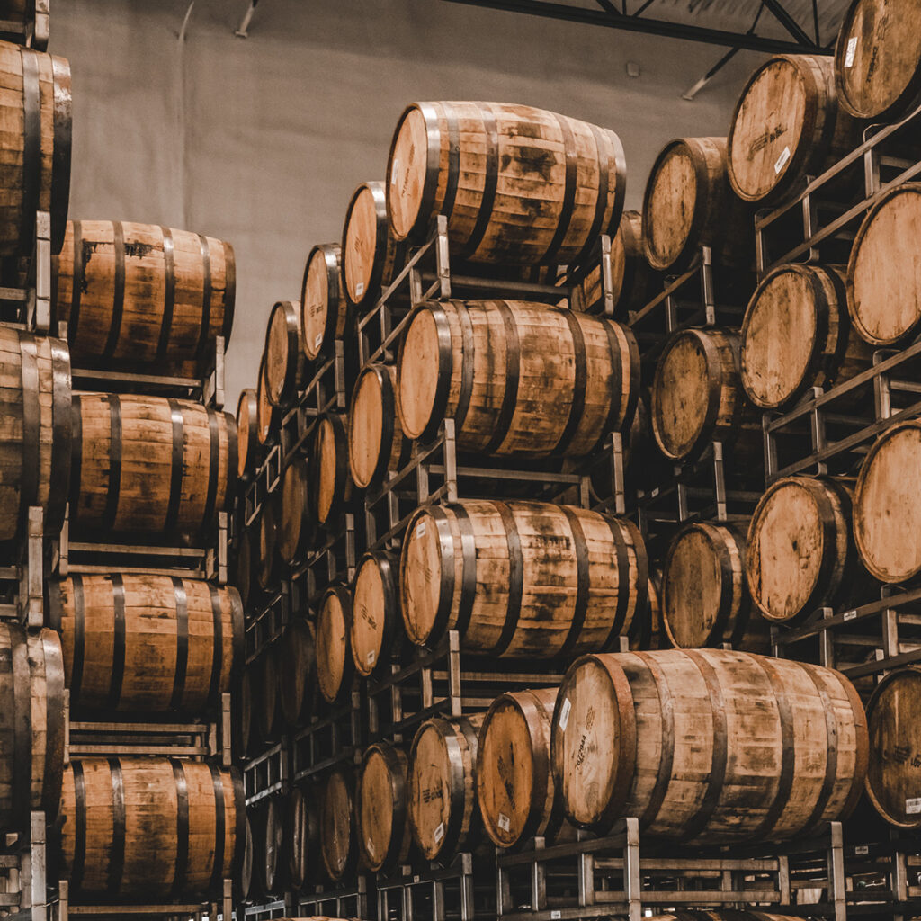 A storage area filled with rows of large wooden barrels stacked on metal racks. The barrels are organized in multiple layers, creating a structured and neat arrangement. The surrounding environment has an industrial feel with high ceilings and overhead lighting.