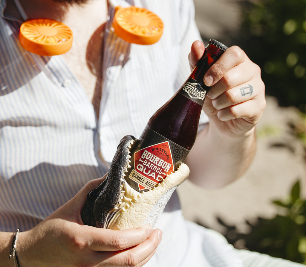 A person with an orange neck fan is holding a bottle of bourbon barrel-aged beer using an alligator-shaped bottle opener. The person is wearing a striped shirt and has a small tattoo on their index finger. The background is outdoors with green foliage and sunlight.