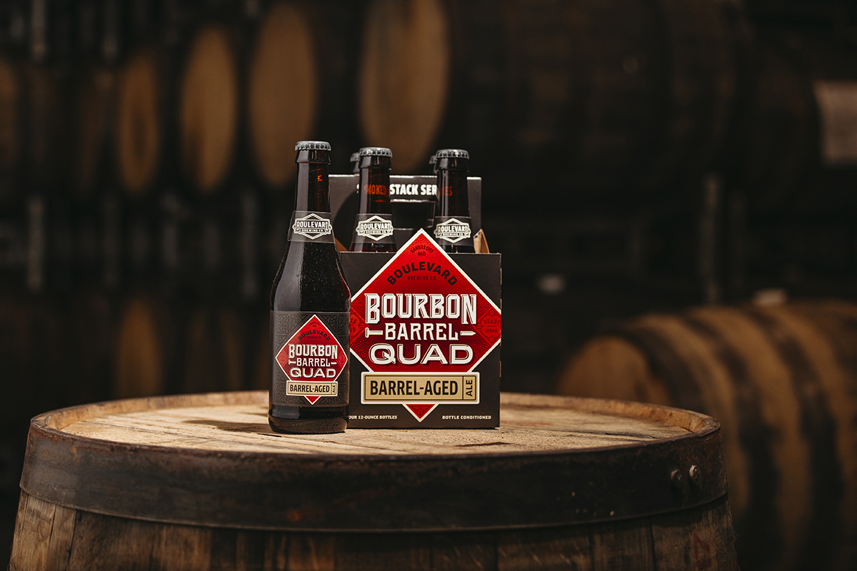 Four bottles of Boulevard Bourbon Barrel Quad Barrel-Aged beer sit in a branded cardboard carrier on top of a wooden barrel. The background features more barrels, suggesting a brewery or cellar setting. The bottles and packaging prominently display the product's name and design.