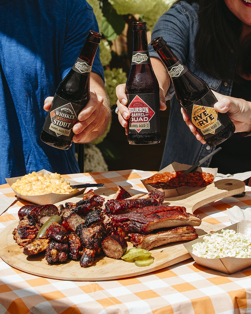 Three people holding different craft beer bottles over a picnic table with a checkered tablecloth. The table has a wooden tray filled with barbequed ribs, sausages, and pickles, along with dishes of macaroni and cheese, coleslaw, and baked beans.