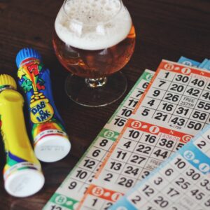 A wooden table with colorful bingo markers, bingo cards, and a glass of beer. The bingo markers are bright with various designs, and the cards display numbers in a grid format commonly used in bingo games.