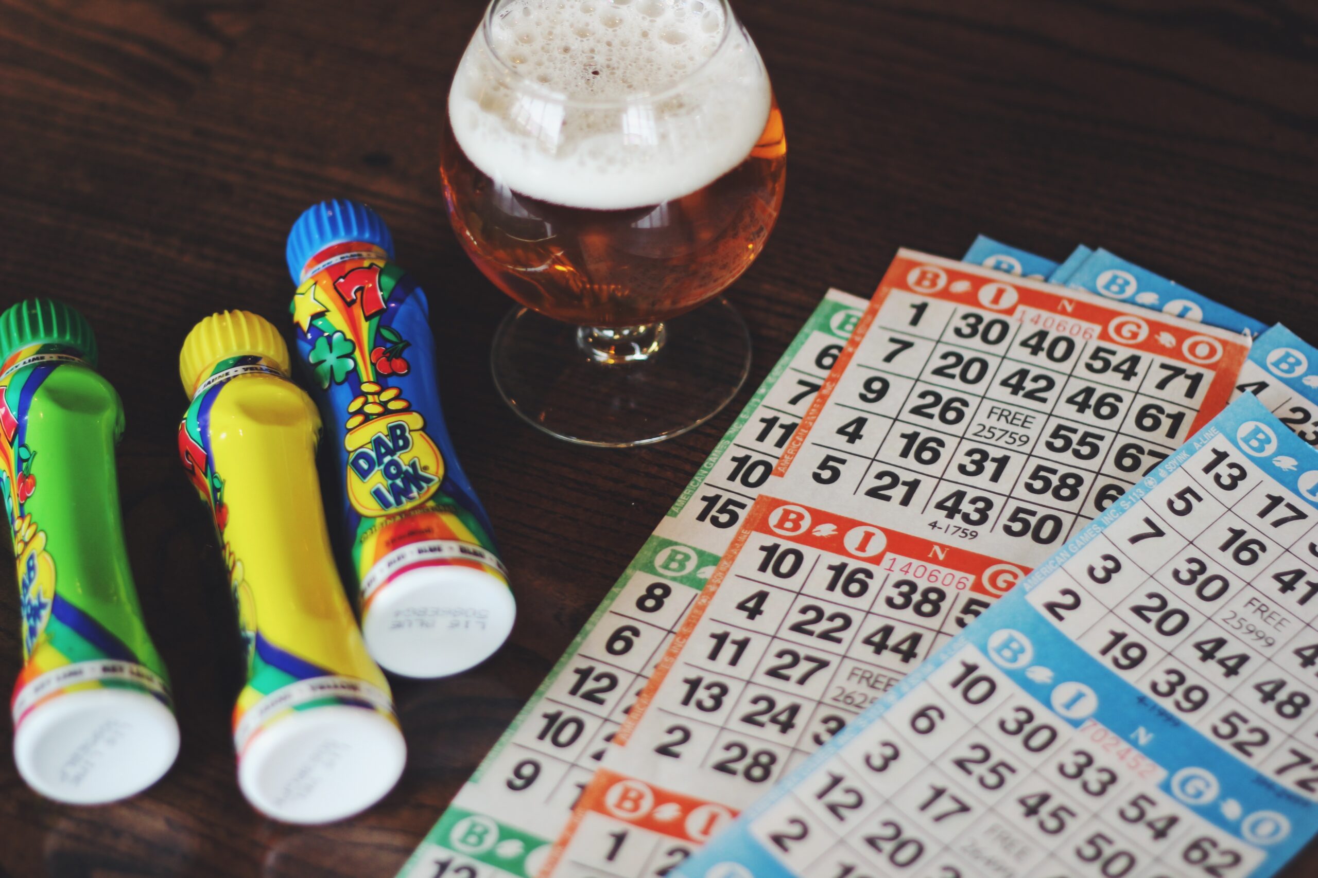 A wooden table with colorful bingo markers, bingo cards, and a glass of beer. The bingo markers are bright with various designs, and the cards display numbers in a grid format commonly used in bingo games.