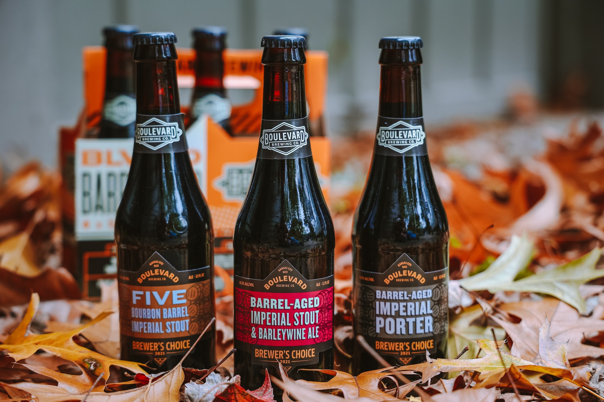 Three Boulevard beer bottles are arranged on the ground covered with autumn leaves in front of a six-pack carrier. They are labeled “Five Bourbon Barrel Imperial Stout,” “Barrel-Aged Imperial Stout & Barleywine Ale,” and “Barrel-Aged Imperial Porter.”.