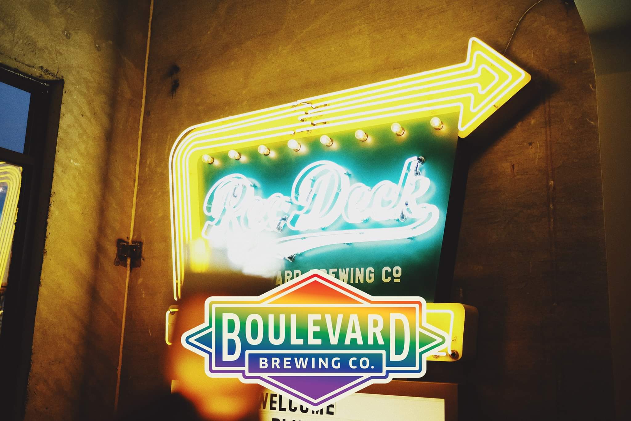 A brightly lit neon sign with an arrow pointing right, reading "Pop Deck" above "Boulevard Brewing Co." The sign features vibrant colors, with the "Boulevard Brewing Co." logo prominently displayed below in a diamond shape with a rainbow gradient.