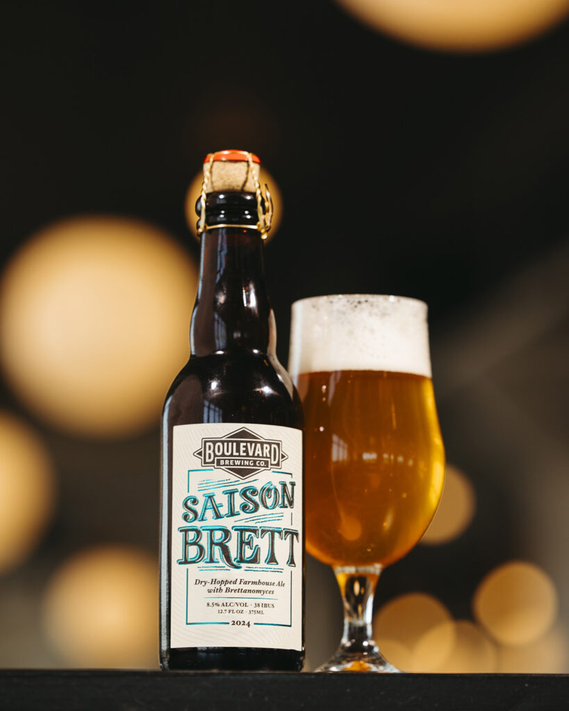 A bottle of Boulevard Brewing Co. Saison Brett stands next to a full glass of the same beer. The label reads "Dry-Hopped Farmhouse Ale." The background is dark with blurred lights creating a cozy ambiance.