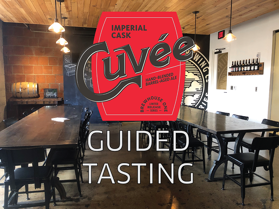 A rustic tasting room with wooden ceiling and floor, featuring long wooden tables and black chairs. A large sign reads "Cuvée Imperial Cask GUIDED TASTING" with details about the hand-blended barrel-aged ale. Shelves with wine bottles are visible on the wall.