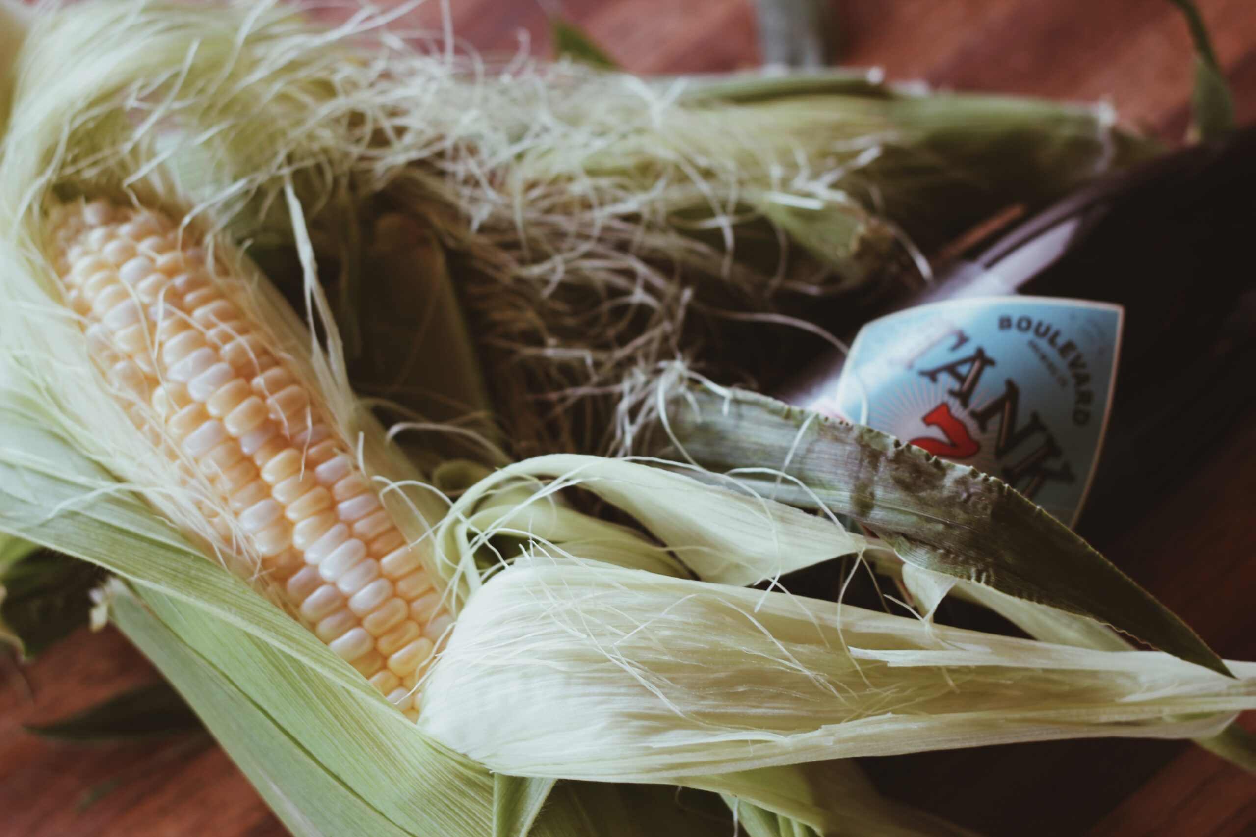 Close-up of a fresh ear of corn with husks partially peeled back, lying next to a bottle with a visible label. The background is a wooden surface.