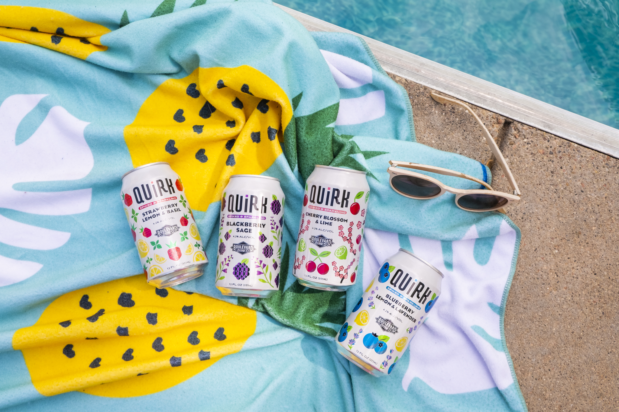 Four cans of Quirk hard seltzer lie on a colorful towel with fruit prints next to sunglasses by a poolside. The cans feature different fruit flavors such as blackberry, lemon basil, and watermelon.