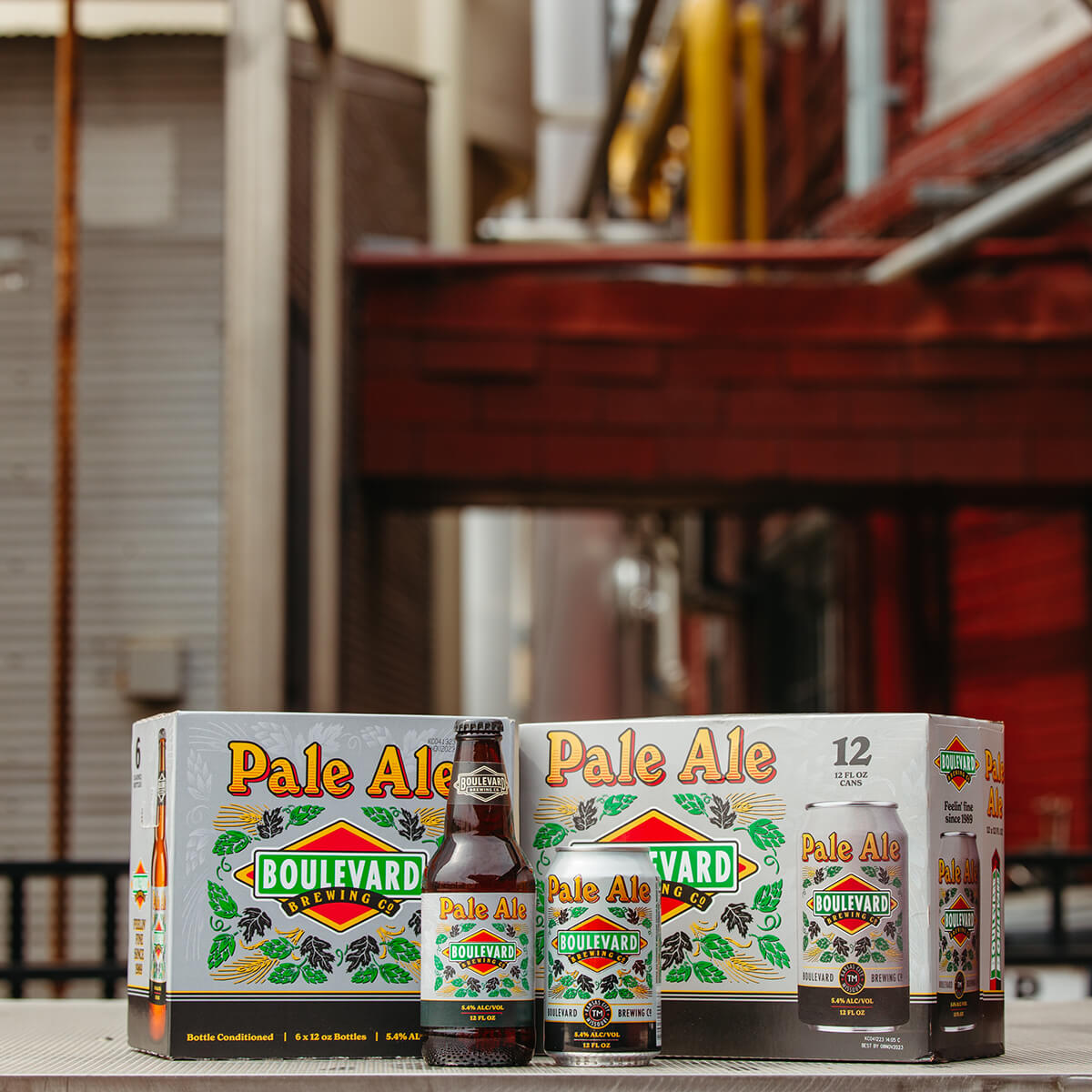 The image shows a 12-pack of Boulevard Brewing Co. Pale Ale beer along with a single bottle and can of the Pale Ale. The packaging is placed on a wooden surface in an outdoor setting with a blurred urban backdrop. The packaging features green hops and orange text.