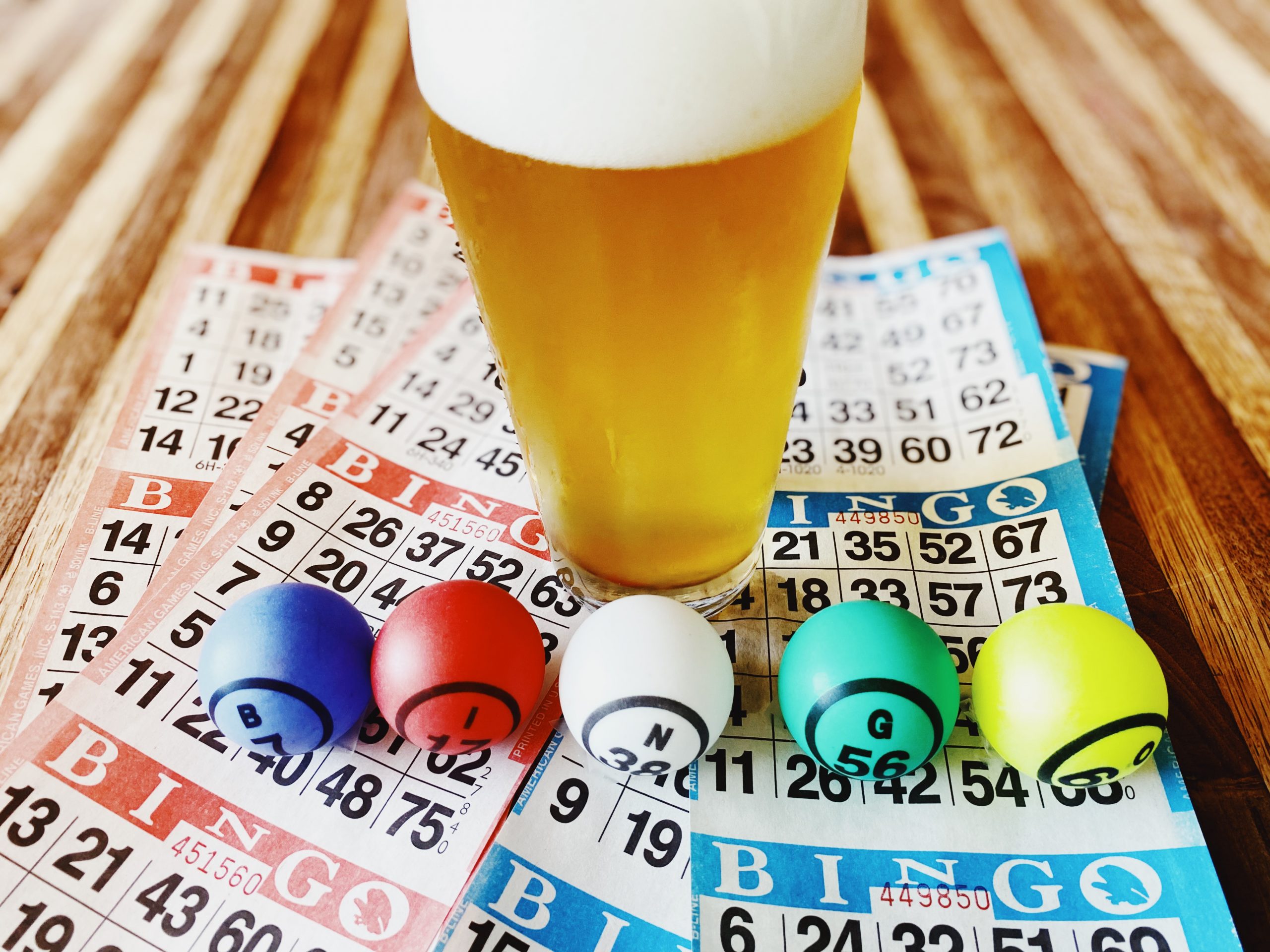 A glass of foamy beer sits on a wooden table surrounded by bingo cards and colorful bingo balls labeled B4, I23, N36, G52, and O61. The cards are a mix of blue, red, and white colors, creating a vibrant and lively scene.