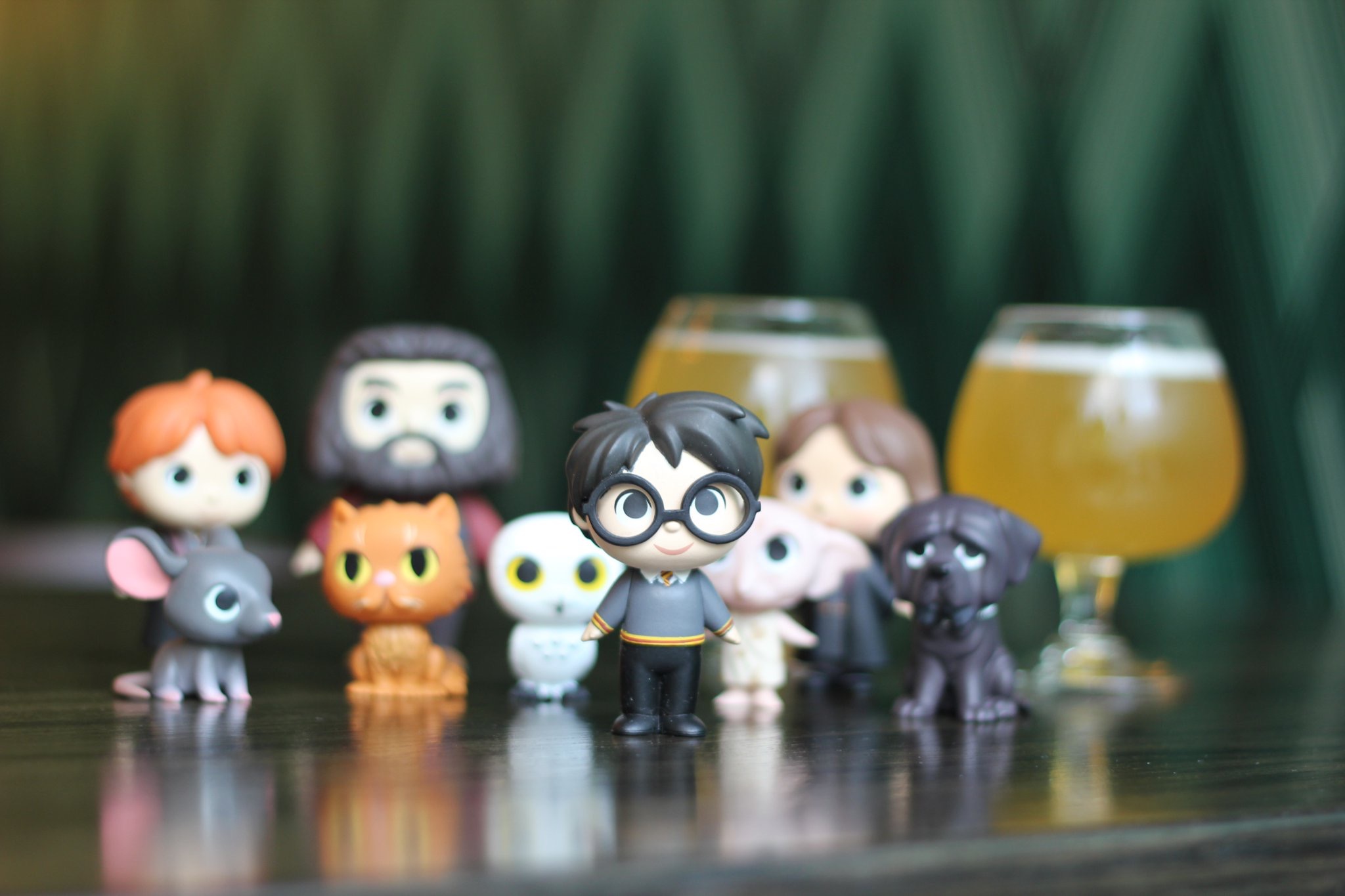 A group of small, stylized figurines resembling characters from a popular fantasy series are arranged on a table. Behind them are two glasses filled with a light beverage. The background is blurred, creating a soft focus effect.