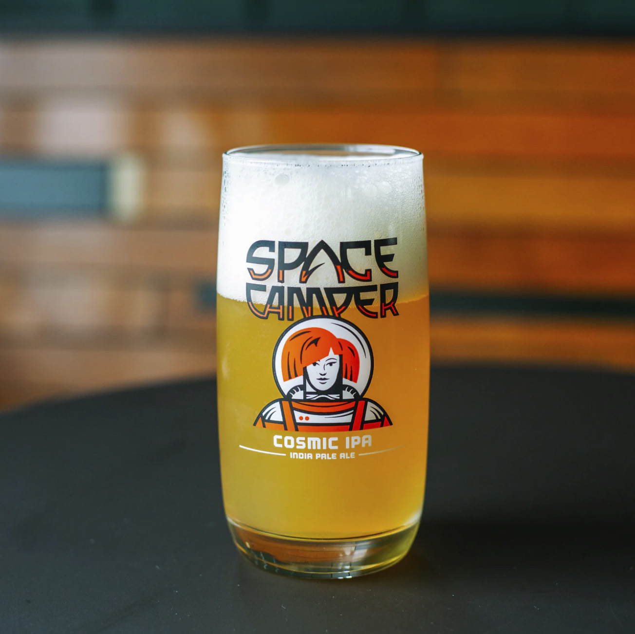 A glass of Space Camper Cosmic IPA beer sits on a dark table, showcasing an illustrated astronaut and the beer's name. This pale golden brew boasts a frothy head, while the softly blurred background adds to its celestial charm.