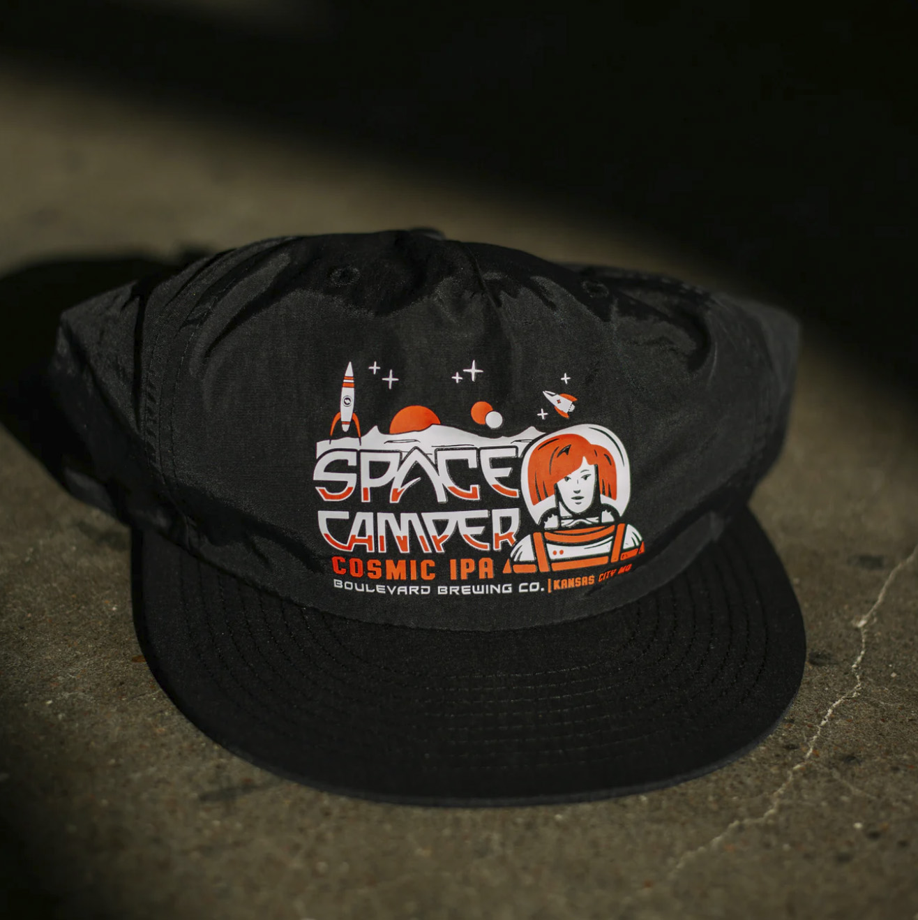 A black baseball cap featuring a colorful space-themed design, perfect for any Space Camper. The front showcases a cartoon astronaut, planets, stars, and the text "Space Camper Cosmic IPA Boulevard Brewing Co." It's set on a concrete surface.