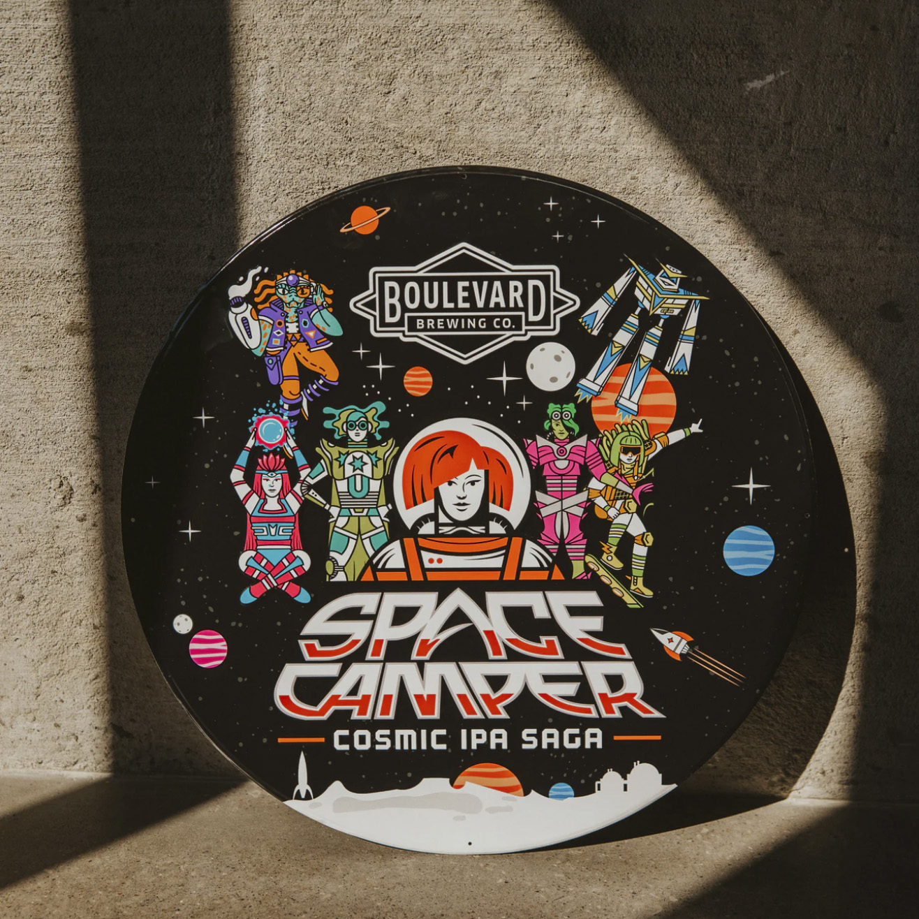 A circular metal tacker for Boulevard Brewing Co.'s "Space Camper Cosmic IPA Saga" is on display, featuring vivid space camper-style characters and planets against a black background, all brilliantly illuminated by sunlight streaming through.