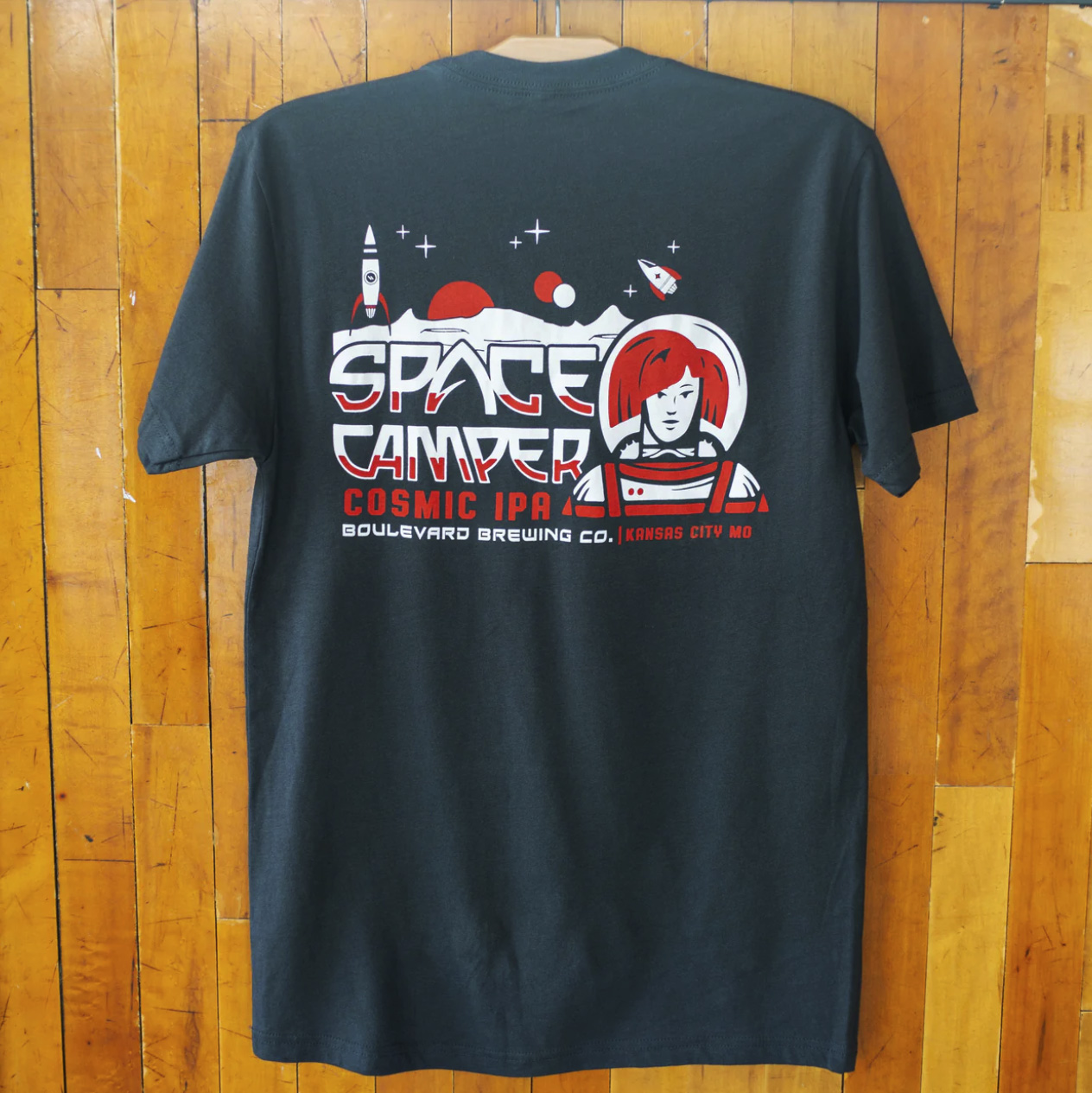 A black T-shirt hangs on a wooden wall, showcasing a space camper design on the back. It features rockets, planets, and an astronaut with the text "Space Camper Cosmic IPA Boulevard Brewing Co. Kansas City MO.