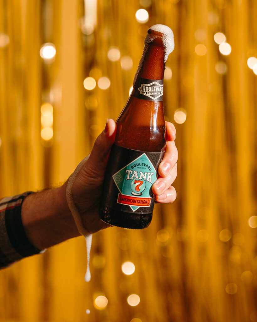 A hand holding a bottle of Boulevard Tank 7 American Saison beer with foam spilling over the top. The background features golden string lights, creating a festive atmosphere.