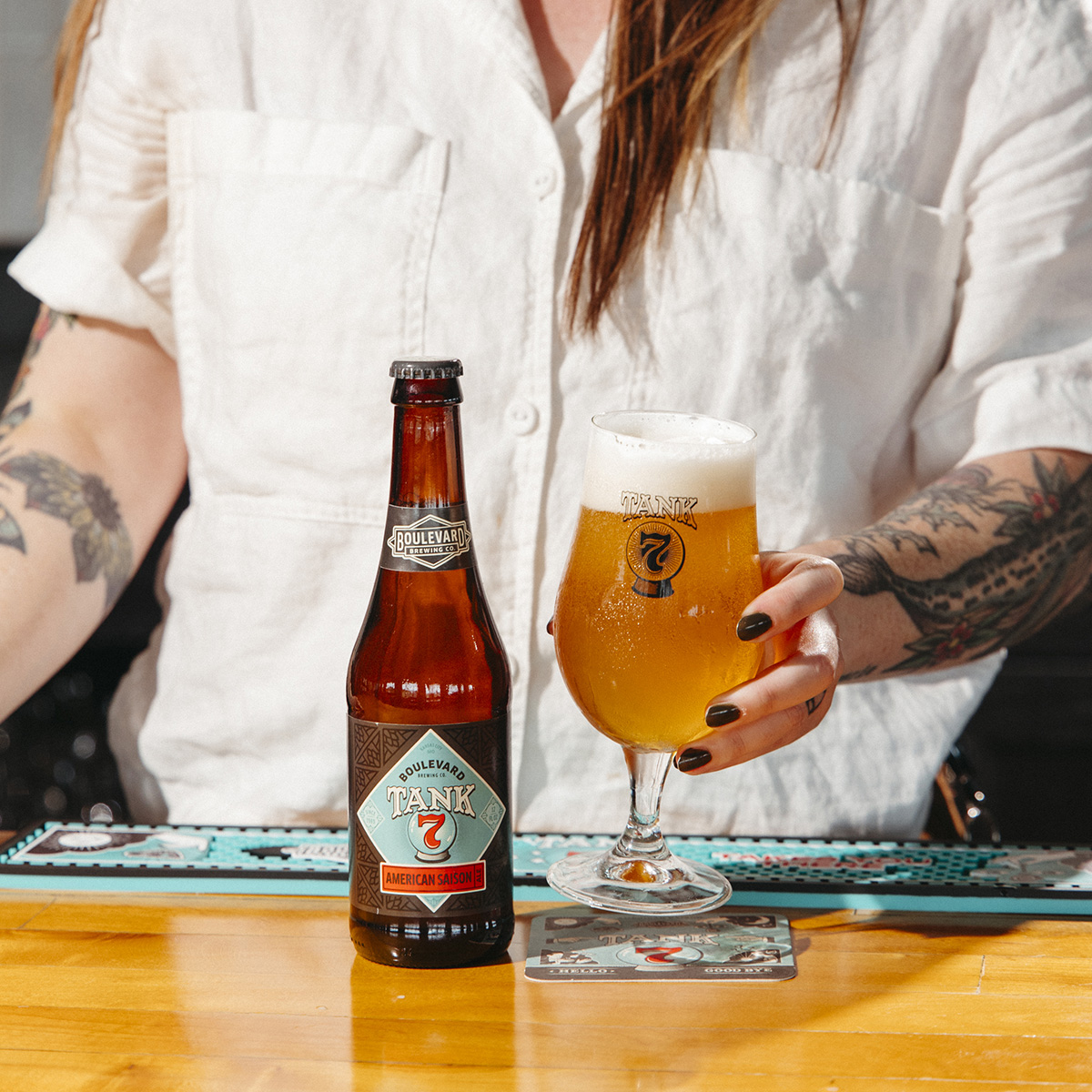 A person with tattoos, wearing a white shirt, holds a glass of Boulevard Tank 7 beer on a counter. A bottle of the same beer is placed next to it. The scene suggests a casual bar or brewery setting.