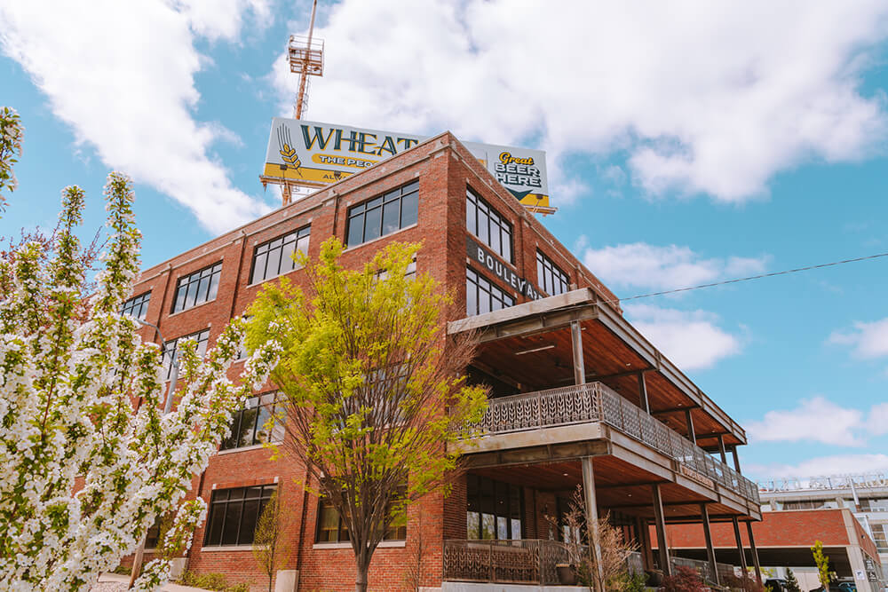 A large red-brick building with multiple windows is adorned with a prominent rooftop billboard advertising "Wheat State," under partly cloudy skies. A tree with green foliage and blossoming white flowers is visible in the foreground.