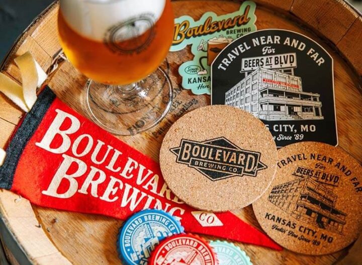 A variety of Boulevard Brewing Co. merchandise is displayed on a wooden surface, including coasters, postcards, a sticker, and a red pennant with the brewery's logo. A glass of beer with the Boulevard Brewing Co. logo sits in the upper left corner.