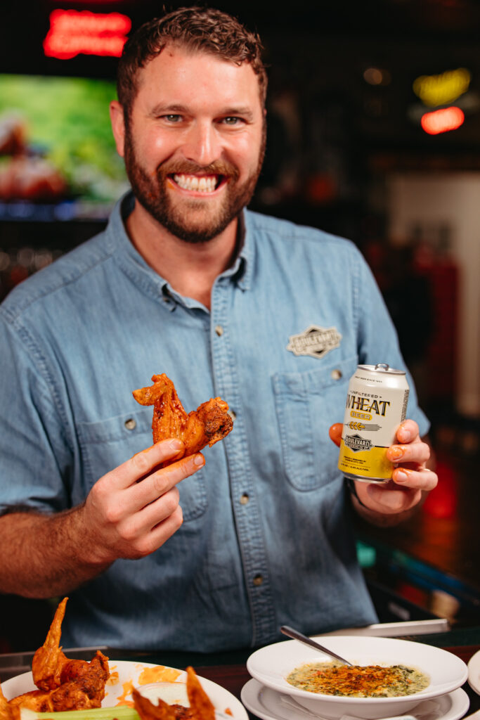 A bearded man with a big smile holds a chicken wing in one hand and a can of wheat beer in the other. He is seated at a table with more chicken wings and a dish of dip. The background indicates a casual restaurant setting with a TV and neon signs.