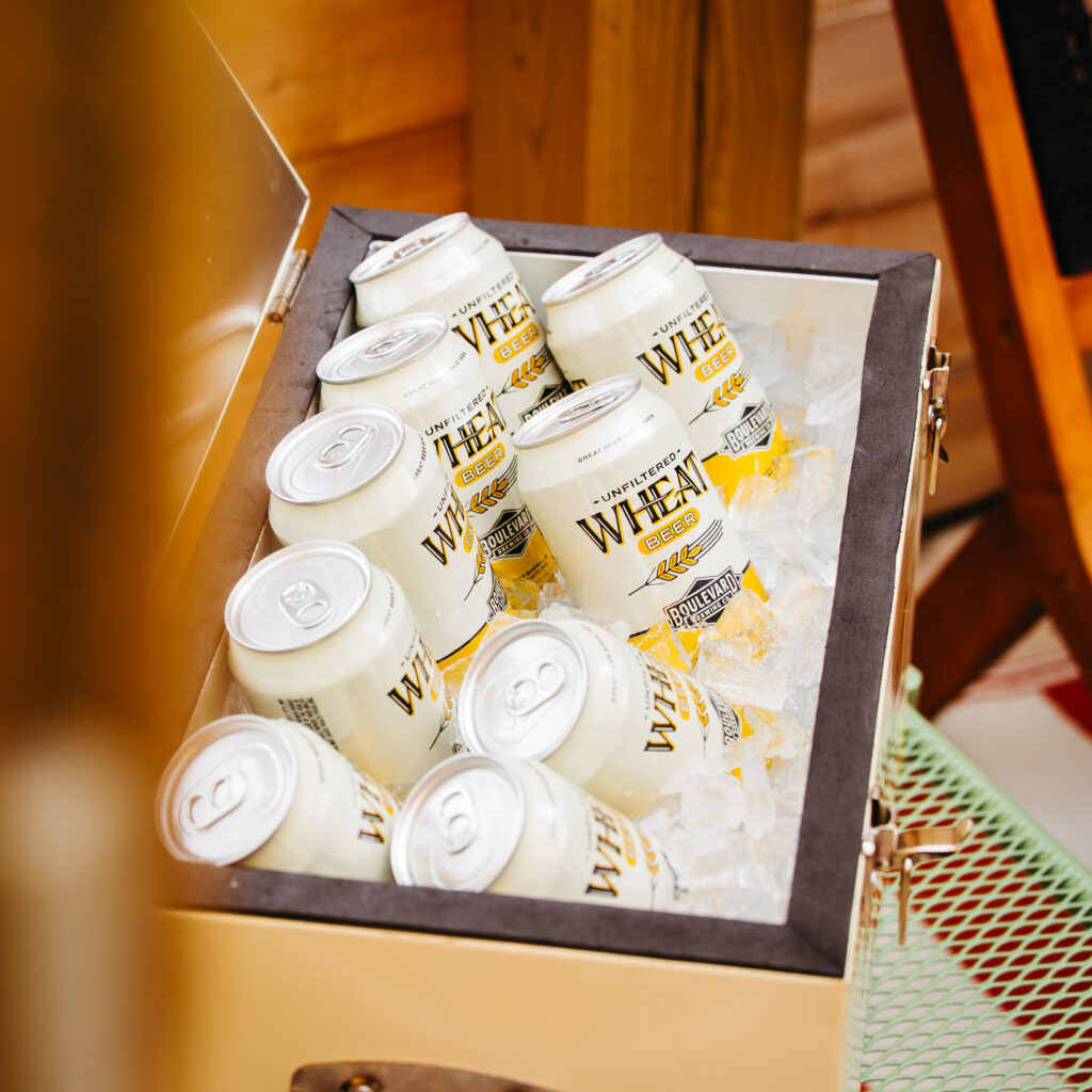 A beige cooler filled with ice and cans of wheat beer. The cans are white with yellow accents and the word "Wheat" prominently displayed along with a graphic of wheat stalks. The cooler is placed on a green mesh surface next to a wooden structure.