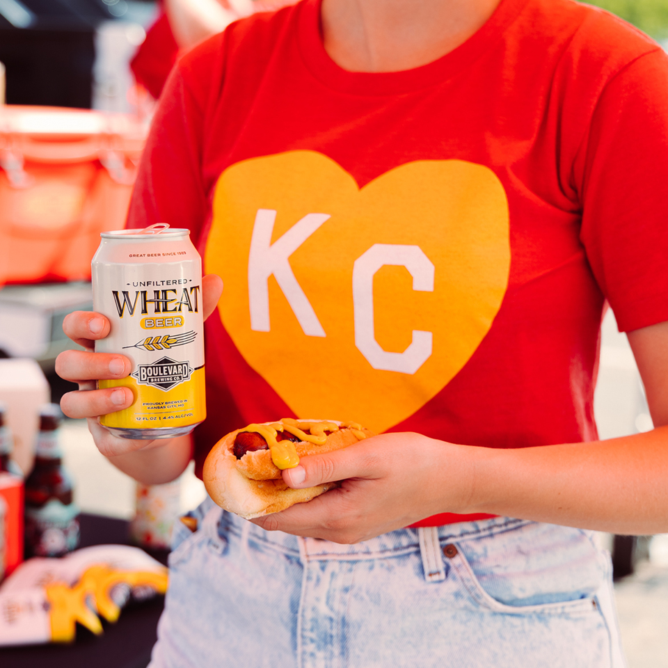 Person holding a can of Boulevard Unfiltered Wheat Beer in their left hand and a hot dog with mustard in their right hand. They are wearing a red T-shirt with a large yellow heart and the letters "KC" in white. The background is slightly blurred and appears to be outdoors.