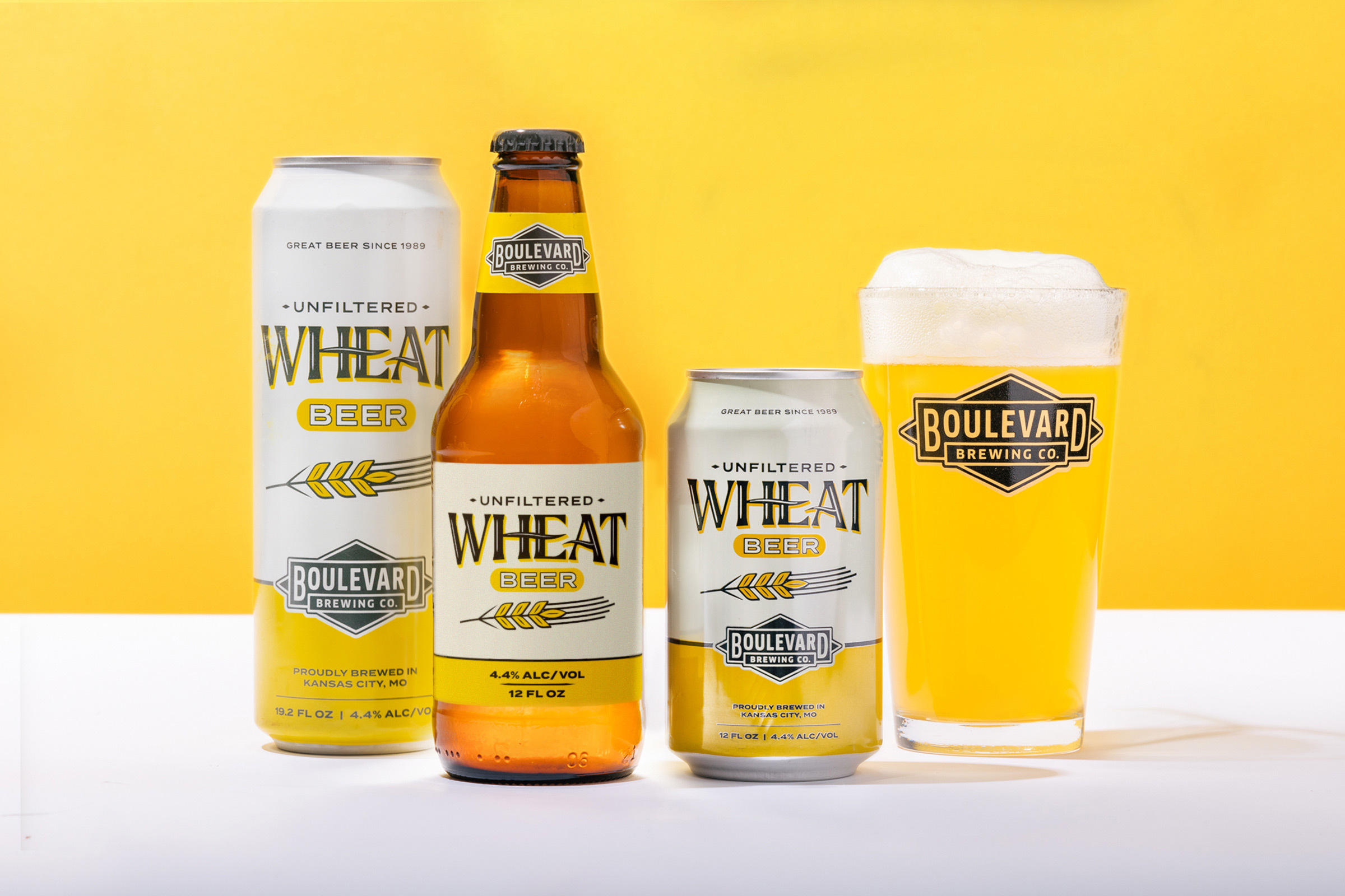 An arrangement of Boulevard Brewing Co. Unfiltered Wheat Beer against a yellow background. The display includes a tall can, a glass bottle, a short can, and a filled glass with a foamy head, all bearing the brand's label.